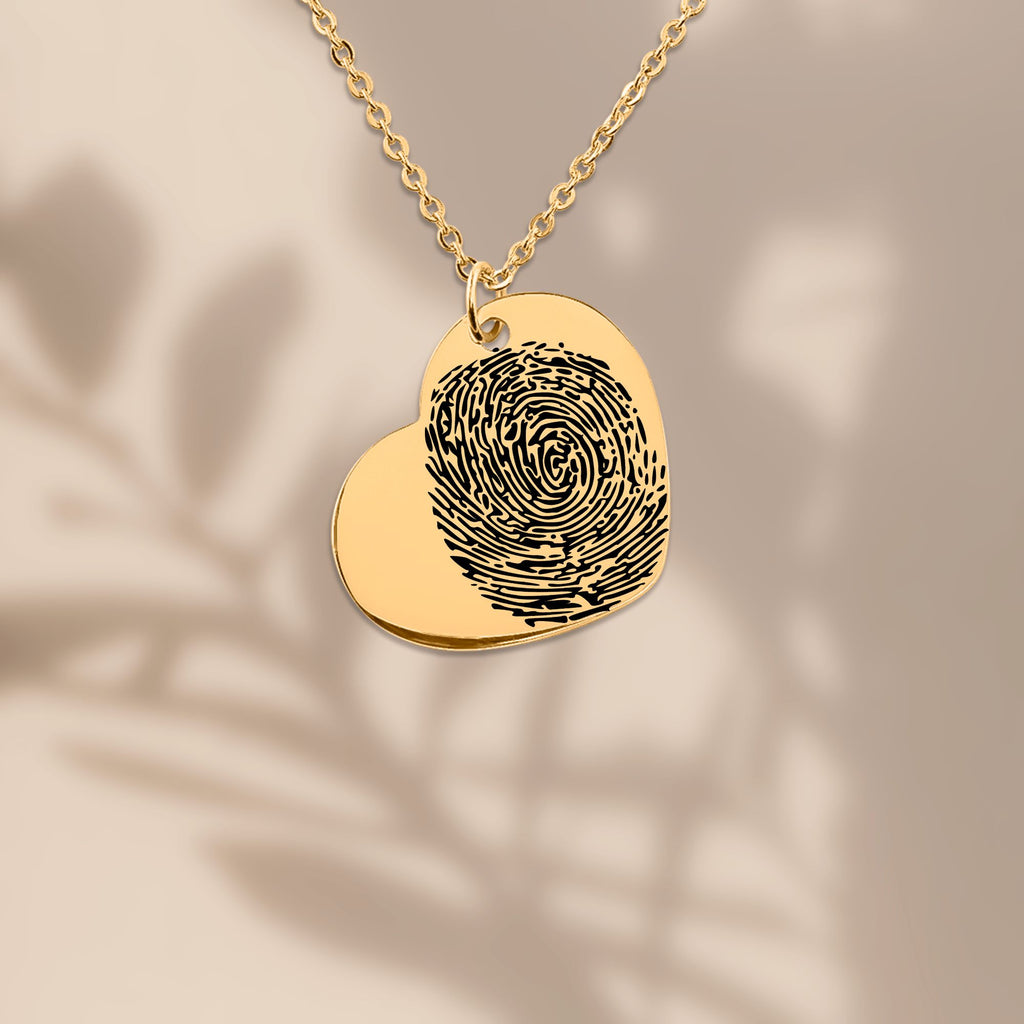 vergoldeter Herzanhänger‑Medallion mit eingraviertem schwarzen Fingerabdruck, hängend an einer feinen kette vor einem warmen, unscharfen Hintergrund