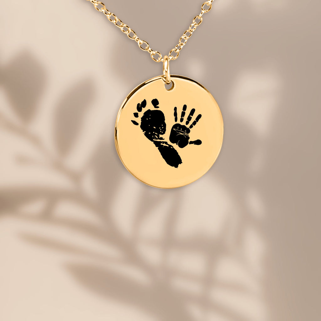 vergoldete Halskette mit rundem, glänzenden Anhänger, auf dem ein schwarzer Babyfußabdruck und ein kleiner Babyhandabdruck zu sehen sind, vor einem beigefarbenen Hintergrund mit weichen Pflanzenschatten