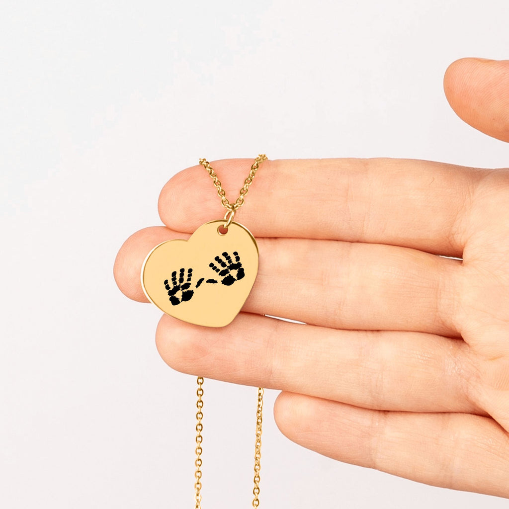 Herzförmiger Anhänger mit zwei eingravierten Handabdrücken an einer vergoldeten Kette, in einer Hand gehalten als personalisierter Erinnerungsschmuck