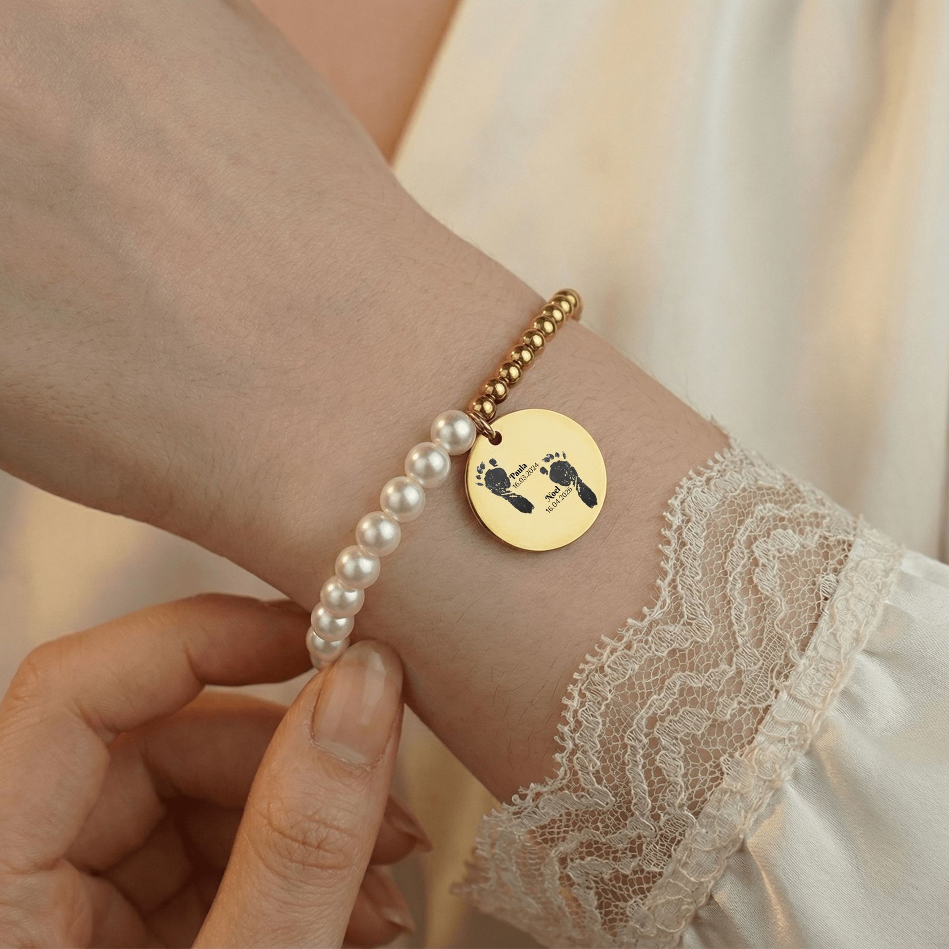 Lifestyle-Foto einer Hand, die eine personalisierte Armband  mit Fußabdrücken, Namen und Geburtsdatum berührt, mit sichtbarem Perlenarmband am Arm