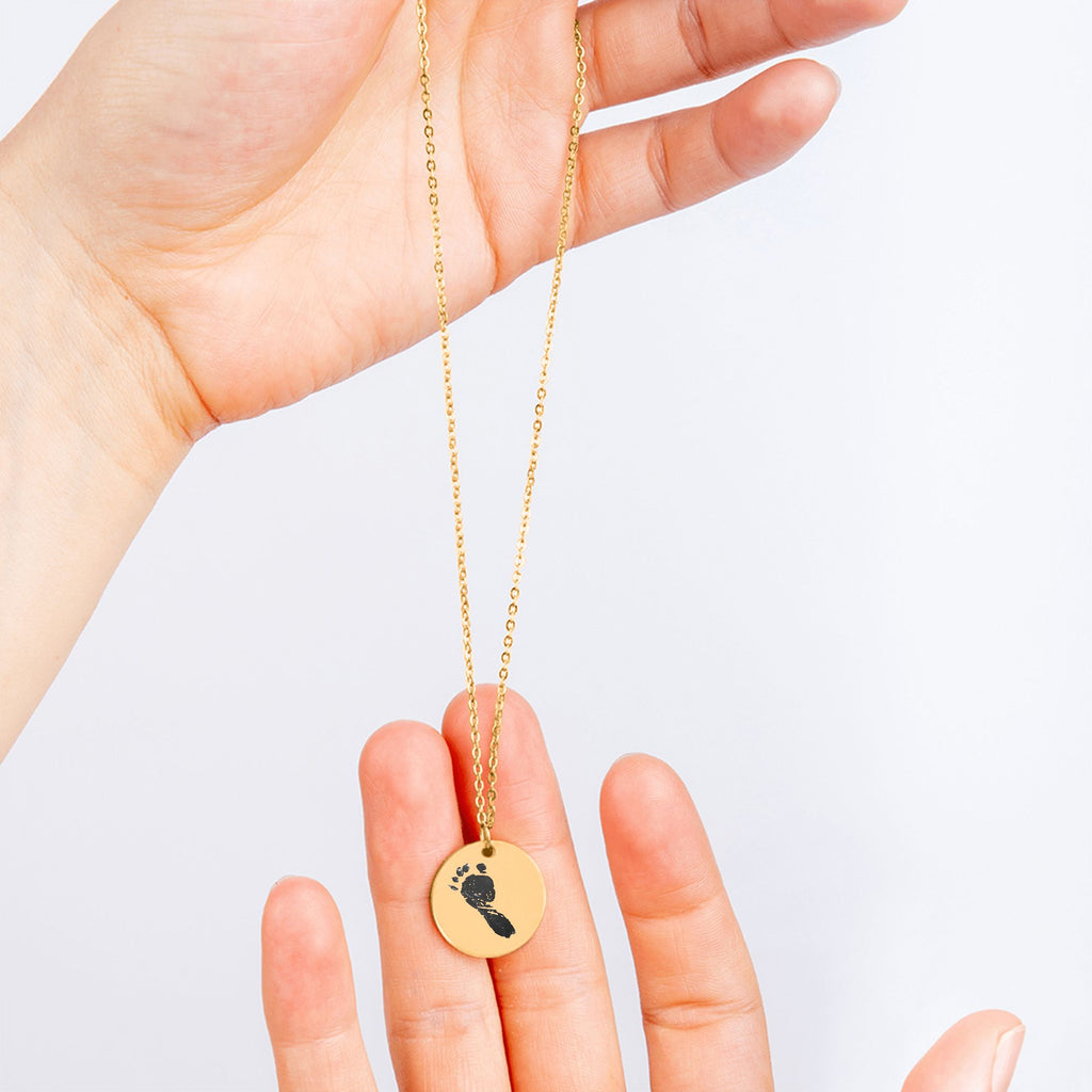 Eine Hand hält eine lange goldene Halskette mit einem runden Anhänger hoch, der einen schwarzen Baby-Fußabdruck als Gravur trägt