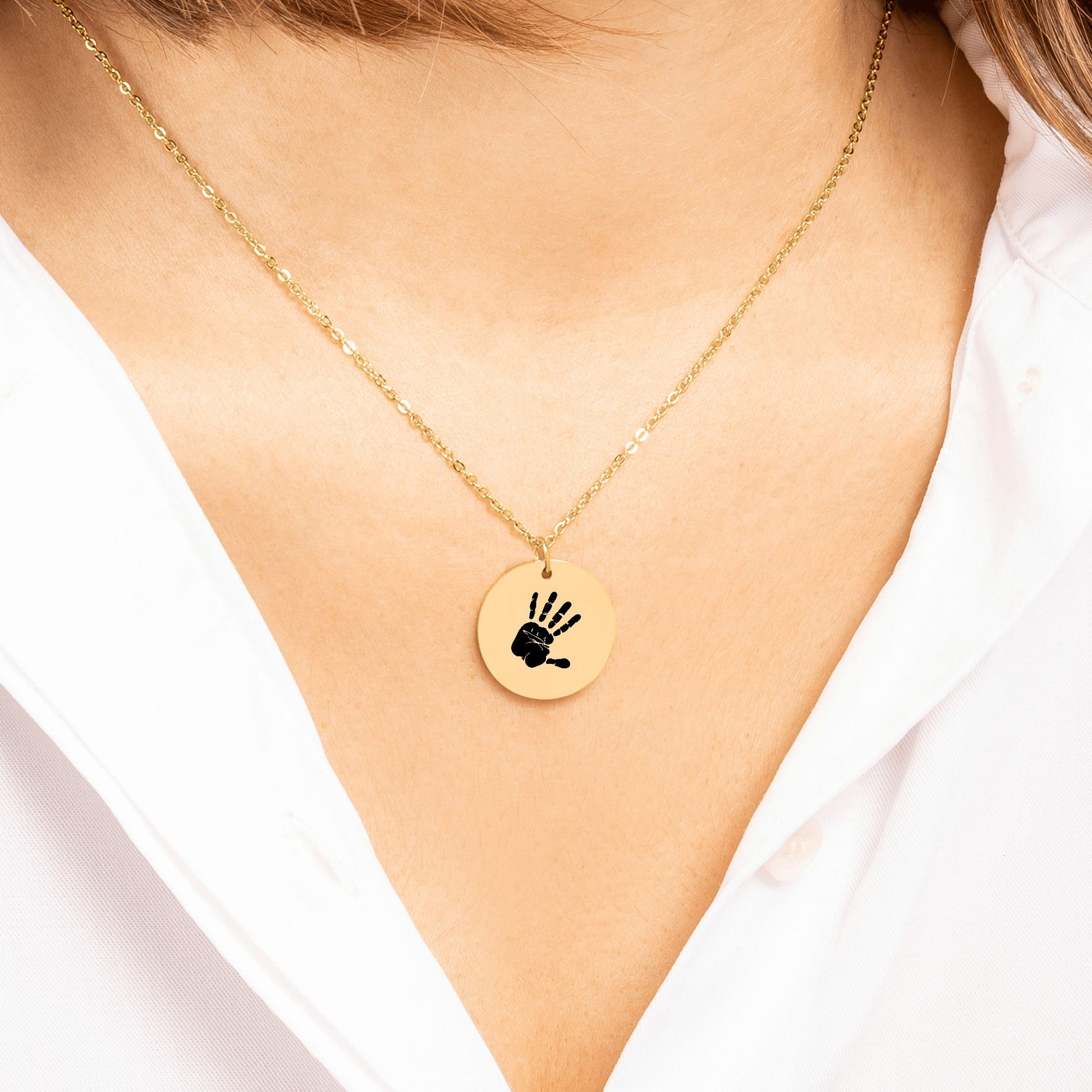 Nahaufnahme einer filigranen Goldkette mit rundem Anhänger, der eine detaillierte schwarze Handabdruck-Gravur zeigt, getragen zu einer weißen Bluse.
