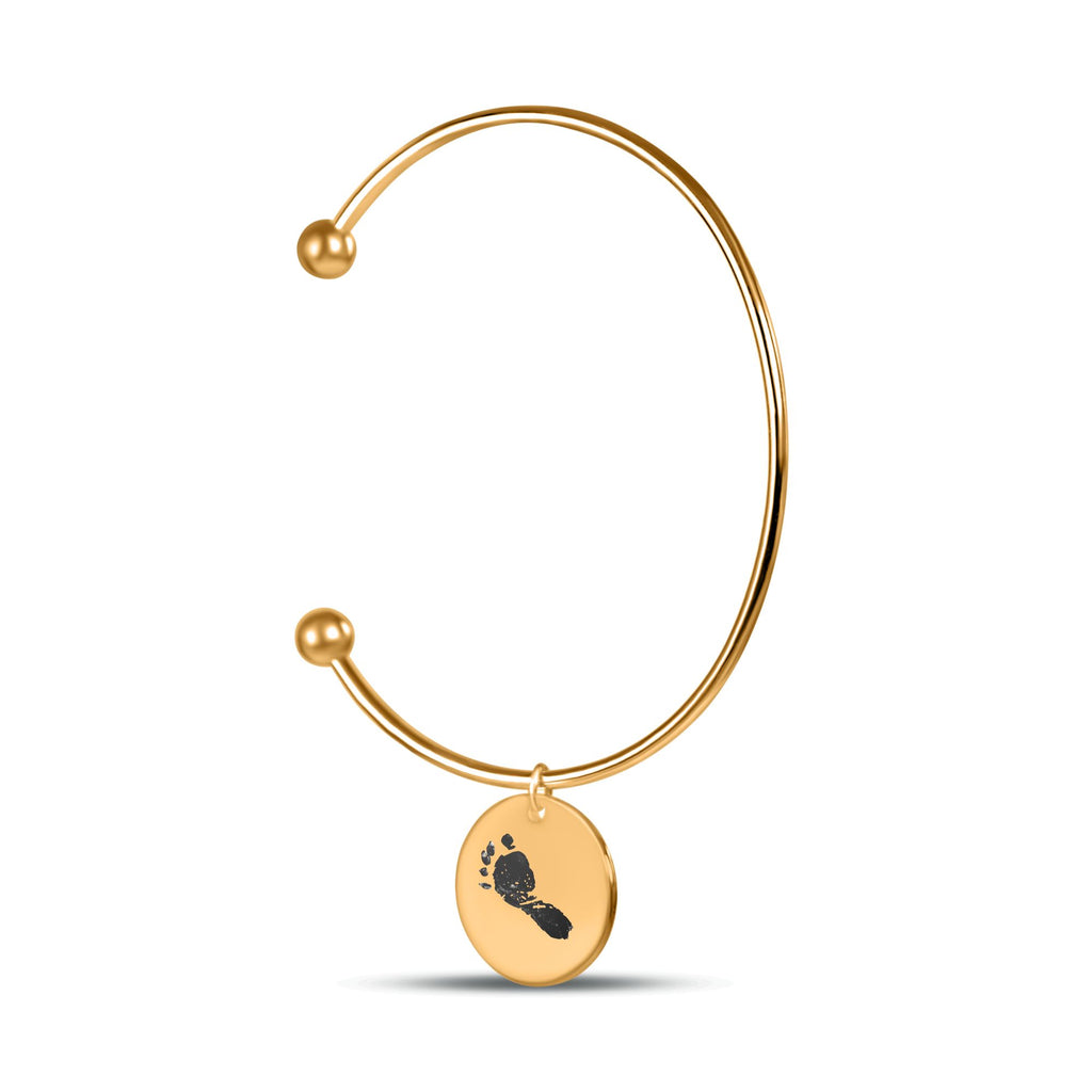 Ein filigraner, glänzend vergoldeter Cuff-Armreif auf weißem Hintergrund. An einer kleinen Öse hängt einen runden Anhänger, die mittig mit einem einzelnen, detaillierten schwarzen Baby-Fußabdruck graviert ist.